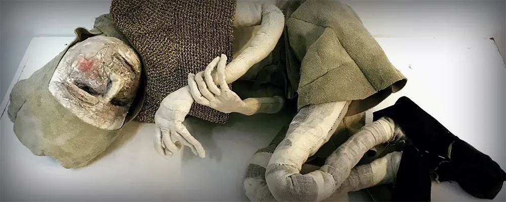 Boy sculpture lying on a plinth dressed in a gold knit shirt, leathery pants, black shoes and a gold-coloured cap.