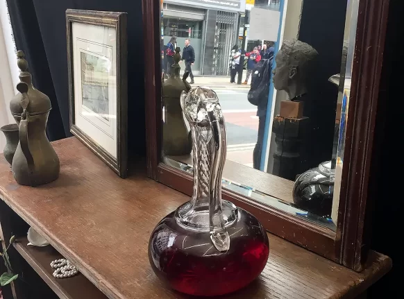 Book shelf with picture frame, a glass flagon with red liquid and a mirror partly reflecting a real life street scene