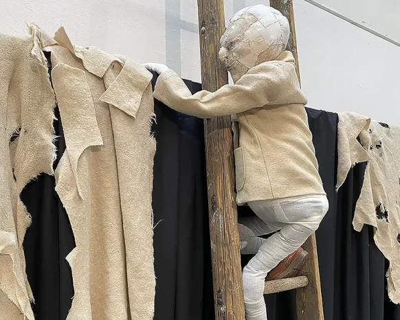 Boy with creamy coloured jacket on ladder climbing up a black curtain with shredded bright fabrics.