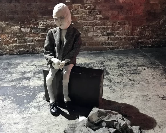 A boy sculpture wearing a tweed jacket sitting on a suitcase with shed fabrics on the floor.