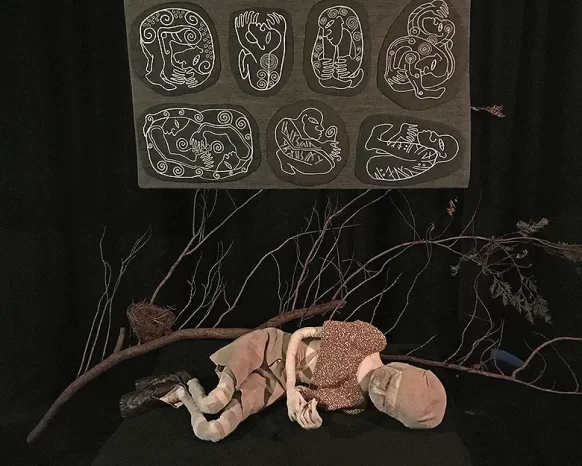 Boy sculpture lying on dark ground in front of a branch and ornamental stitchwork.