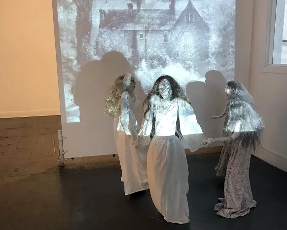 Four girl sculptures in white spooky dresses holding hands and dancing in front of black and white house projection.