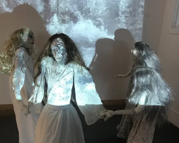 Four girl sculptures in white spooky dresses holding hands and dancing in front of black and white projection.