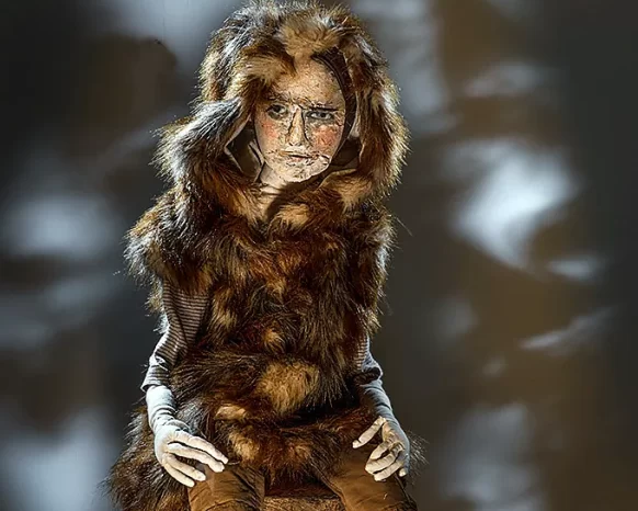 Boy sculpture in fur coat sitting on a tree trunk before a dark background